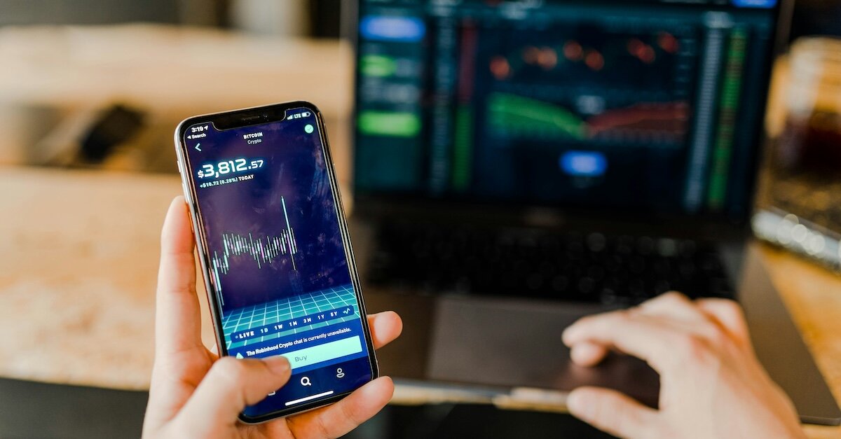 A person looks at stock movement charts on their cell phone, while more detailed charts are on a laptop screen in the background.