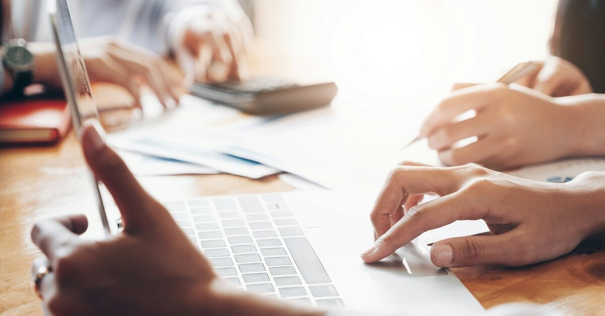 An individual works at a laptop computer while others around the person review a calculator and contracts