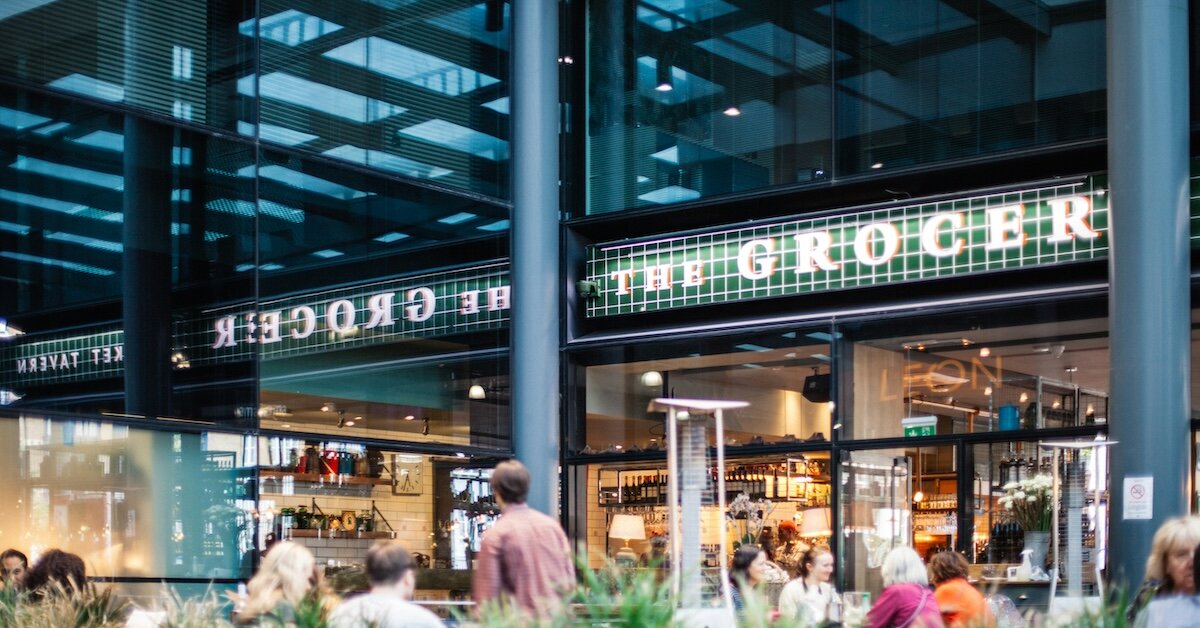 An indoor grocery store with pedestrians walking in front of an outdoor eating area