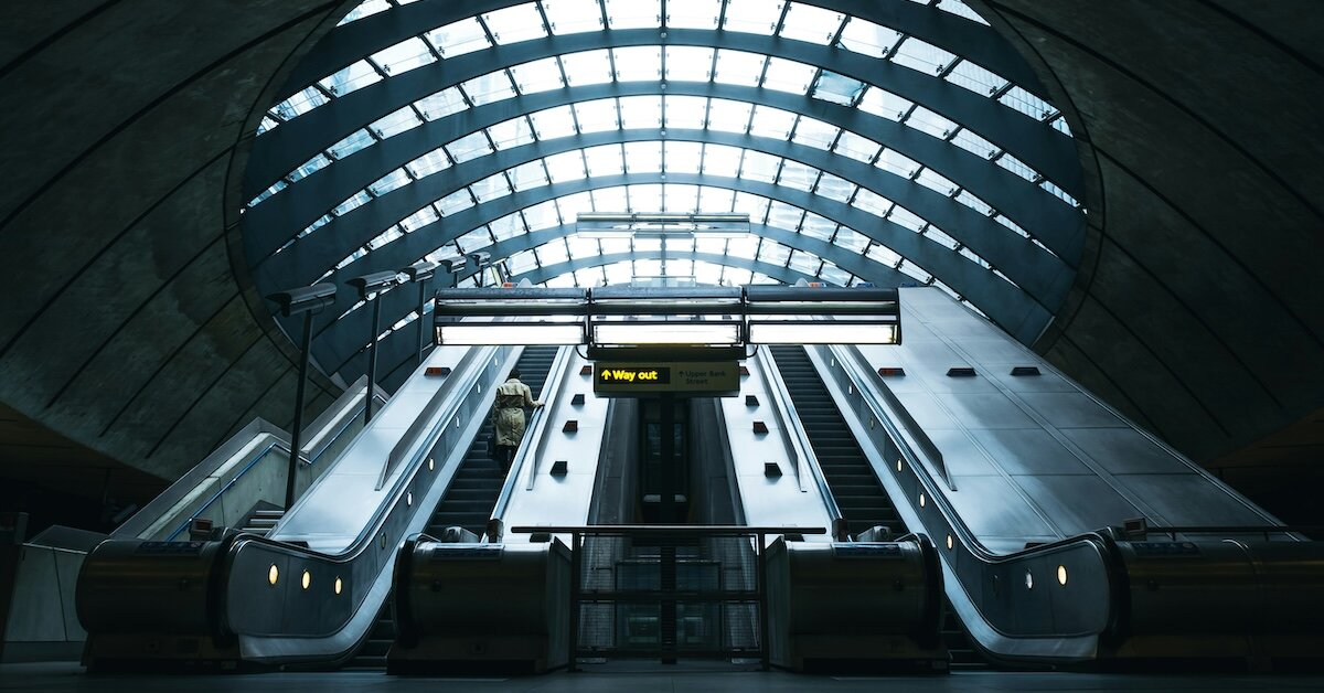 The inside of a train station with escalators and sunlight pouring through overhead glass windows