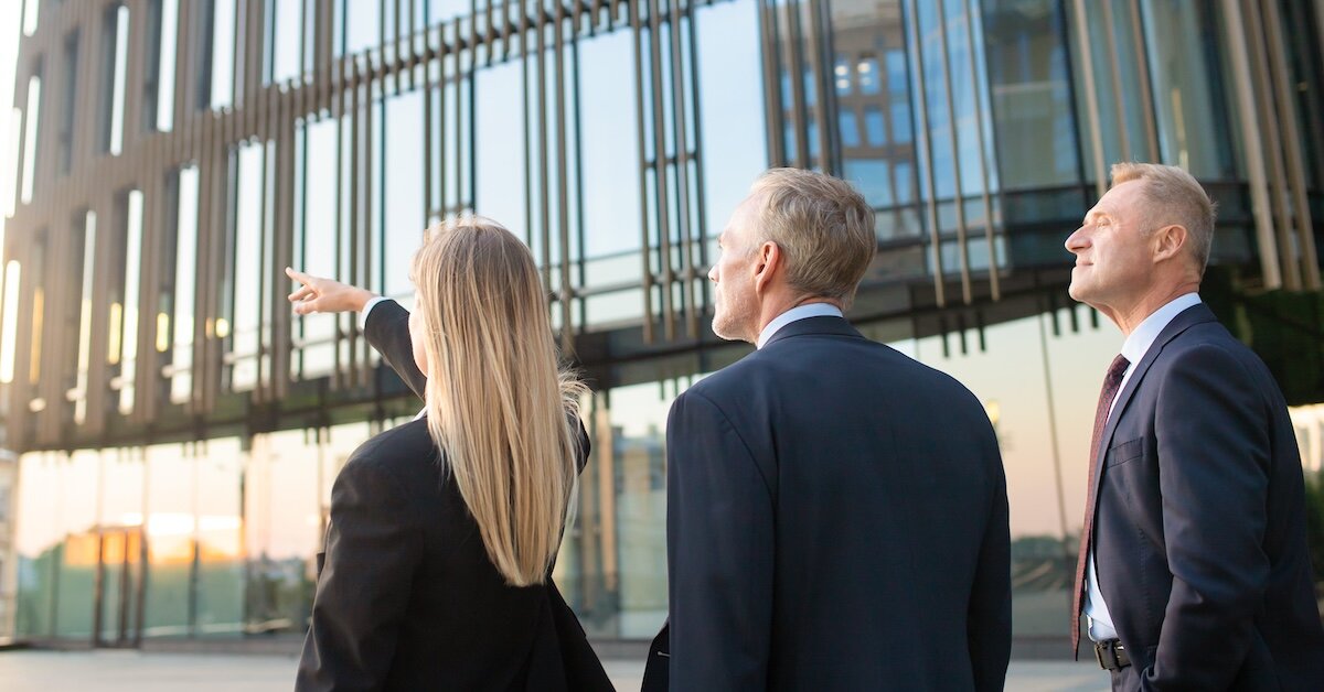 Three business professionals point and look at a commercial building
