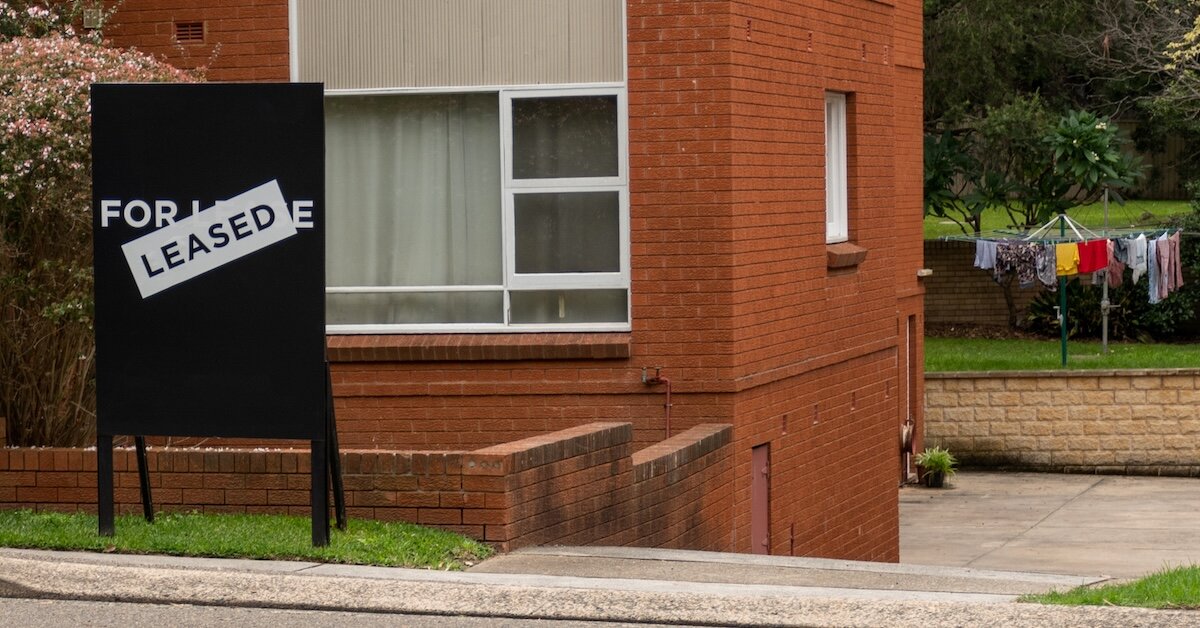 A brick building with a For Rent sign slapped over with a "Leased" sticker in front of it