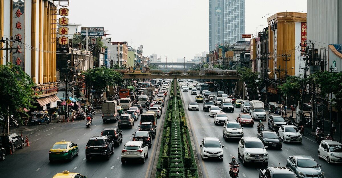 Several cars driving down a road with trees and greenery lining the city streets