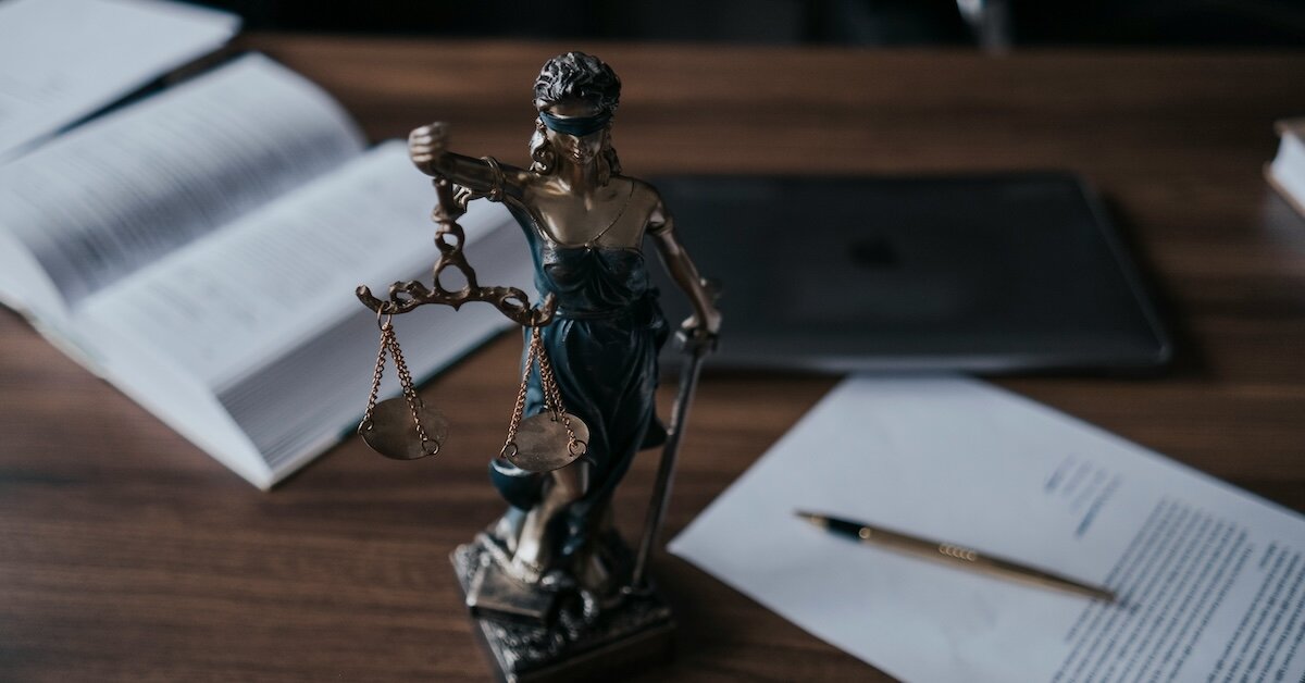 a statue of Justice sits in the foreground on a table with a legal book and contract behind it