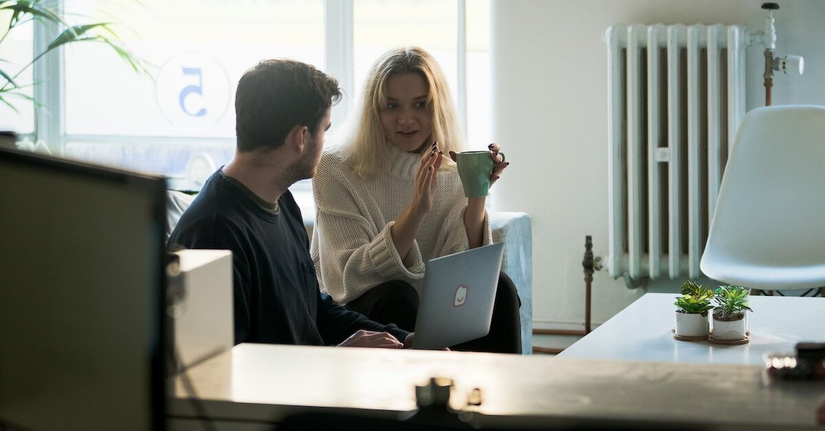 A man and woman discuss a project on a laptop screen nearby a desk in the foreground