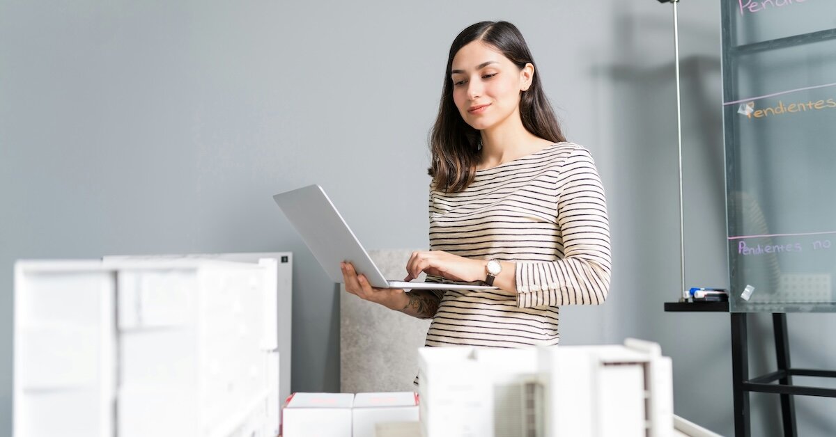 A woman reviews appraisals while standing with her laptop over models of commercial buildings on a table