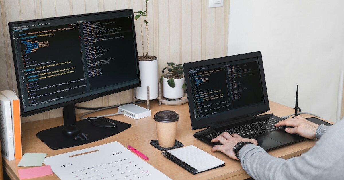 A person coding at their desk computer with a monitor in the back