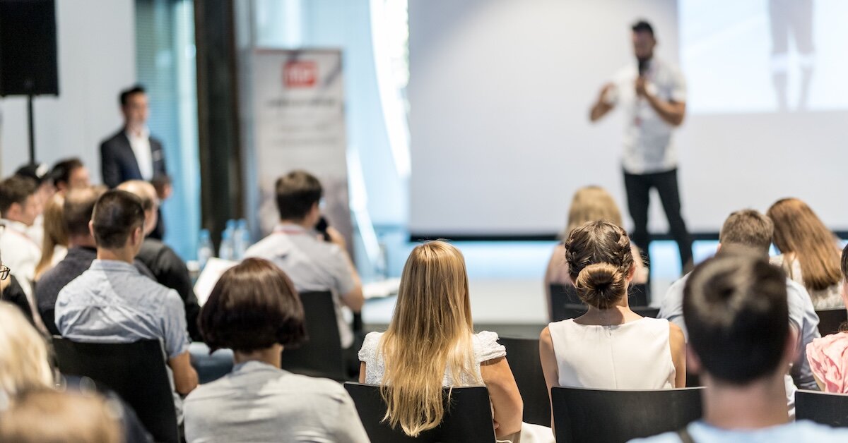 A person presenting on stage to a group of professionals in a conference hall.