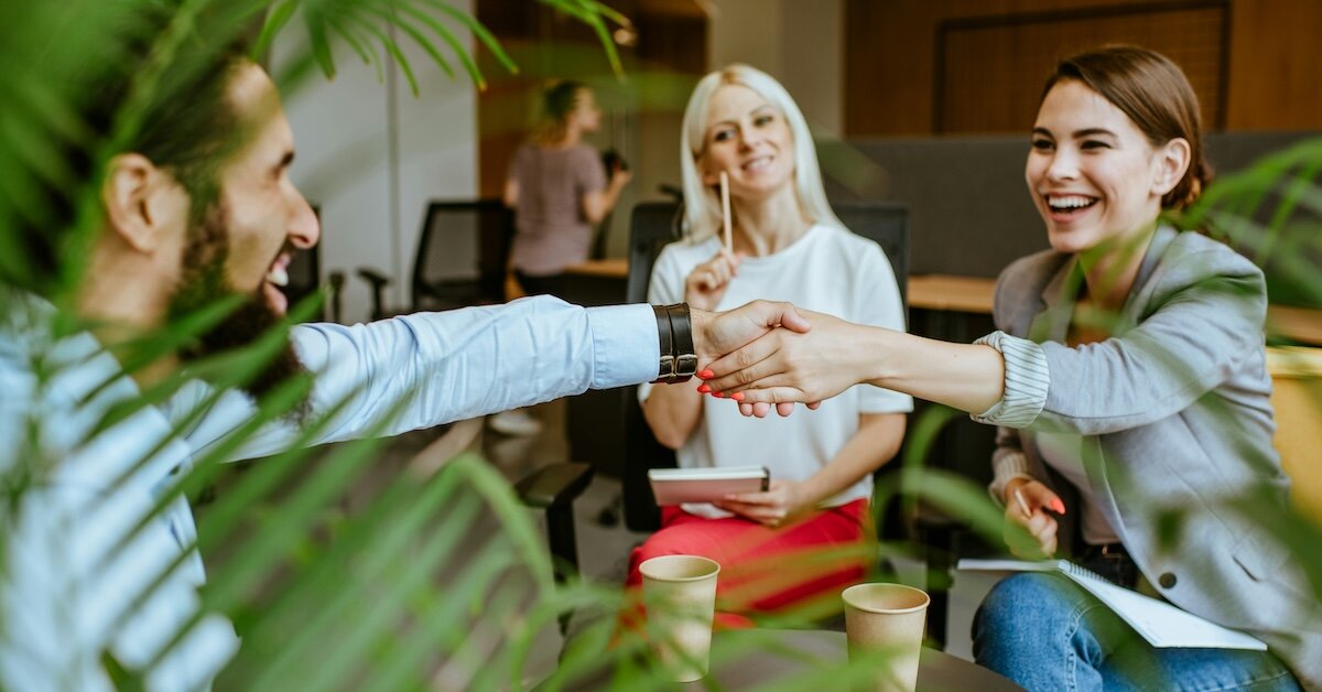 Two people shake hands across a coffee table, with another person looking on holding a notebook and pen in her hand.