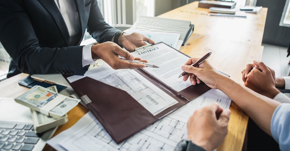 A person signs a contract with stacks of cash and a calculator sitting on top of a desk with architectural blueprints