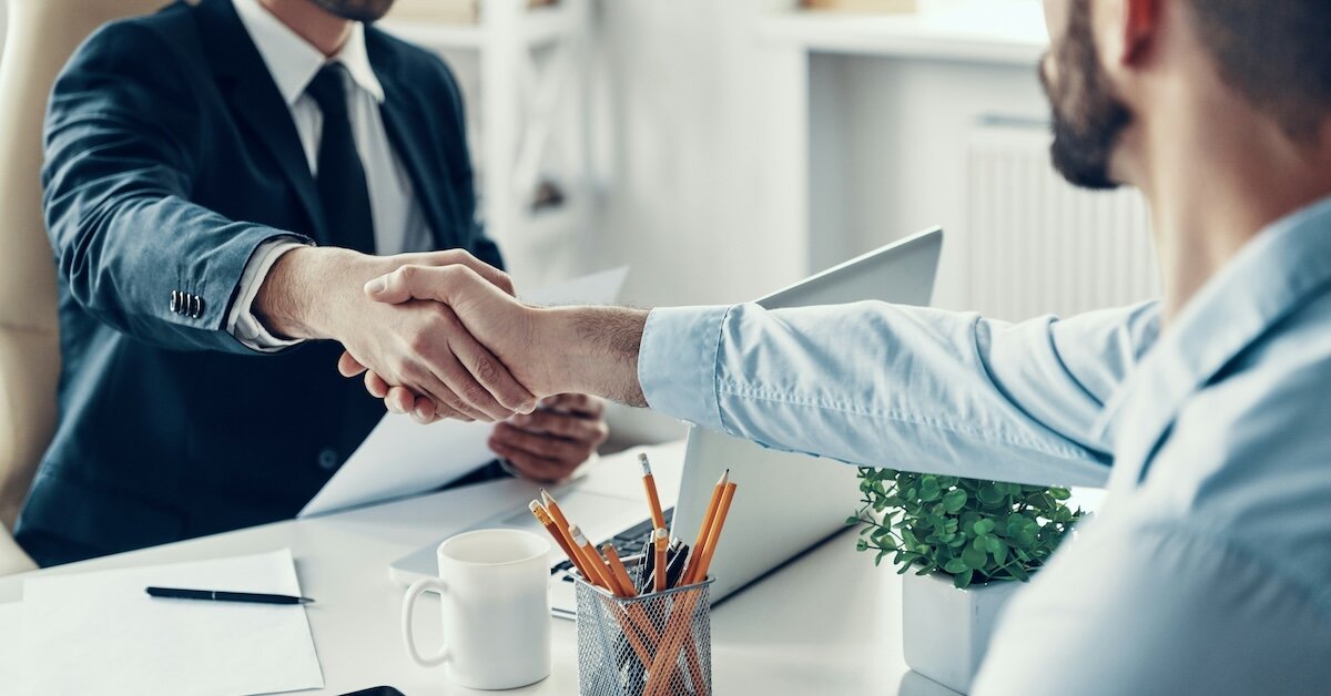 Two young men shake hands across a desk