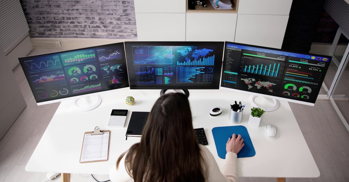 A woman works on a multi-desktop computer set up, reviewing charts and dashboards
