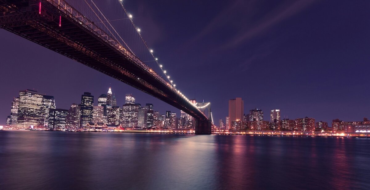 New York City across a bridge at night with the lights reflecting in the water
