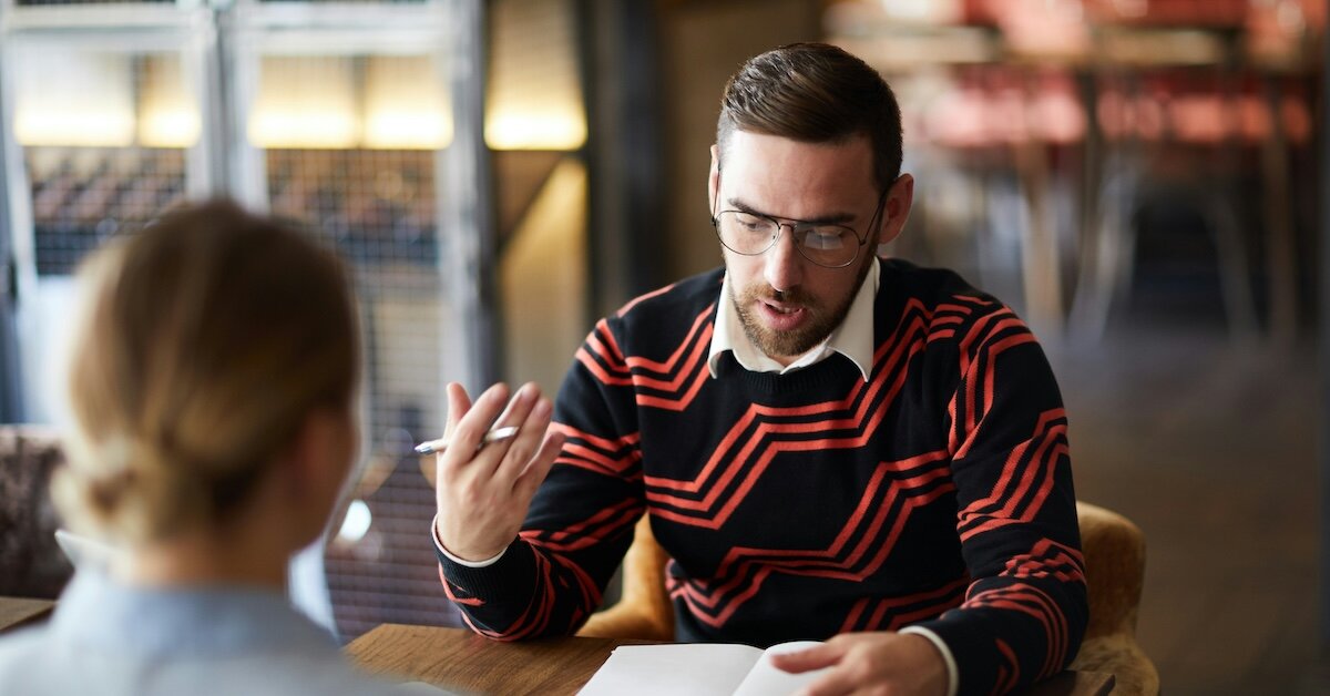two individuals meet at a table in a lobby coffee shop, one holding a notebook
