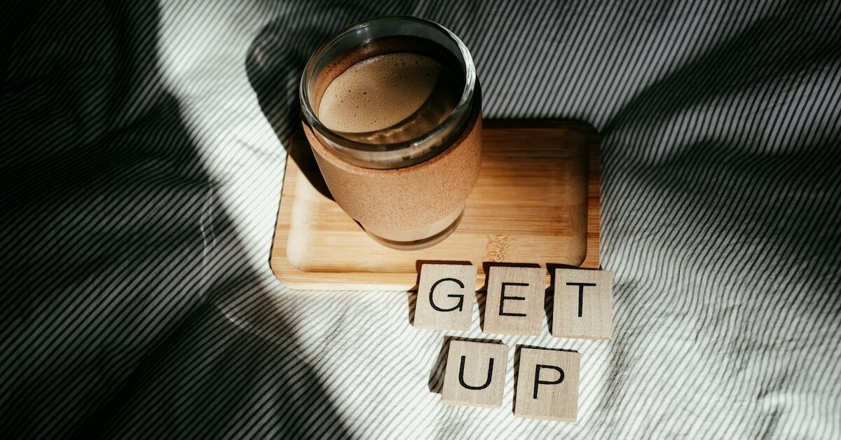 A cup of coffee sits on a wooden tray on a bed, with scrabble tiles spelling out "Get up"