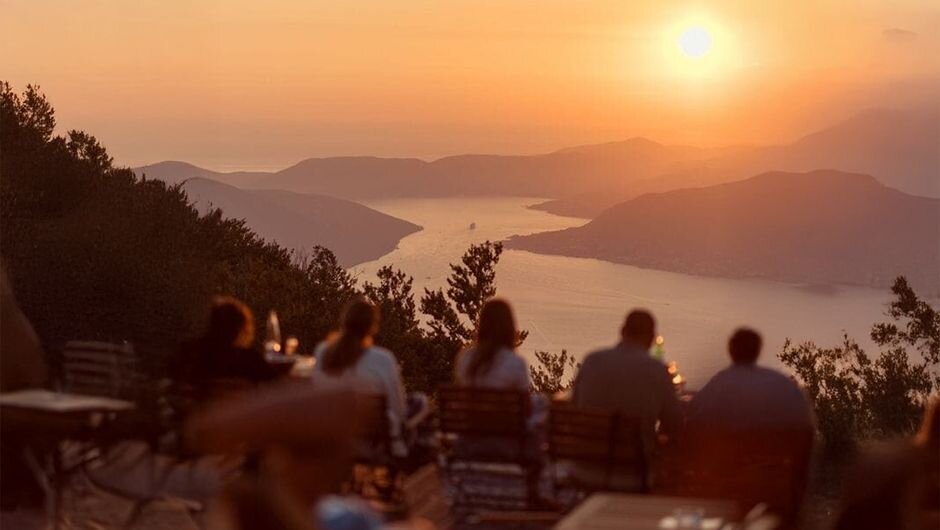 Ljudi sede za stolovima sa pogledom na more i planine, uživajući u prelepom pejzažu.