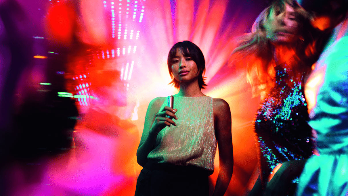 Samouvjerena žena u klubu drži Ploom AURA Lunar Silver, okružena šarenim svjetlima.