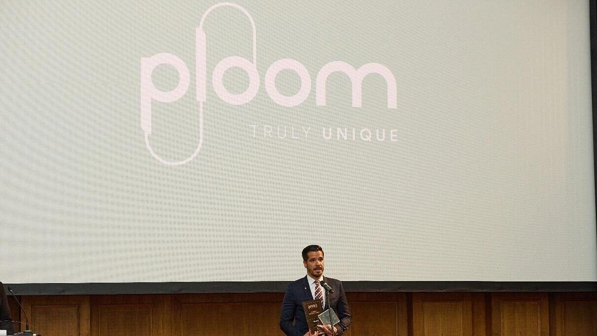 Woman holding a Ploom device while working on a laptop, with a planner and documents on the desk nearby.