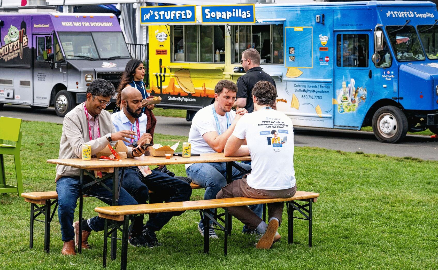 People gathered around colorful food trucks on a sunny day, with customers ordering from green and orange mobile vendors
