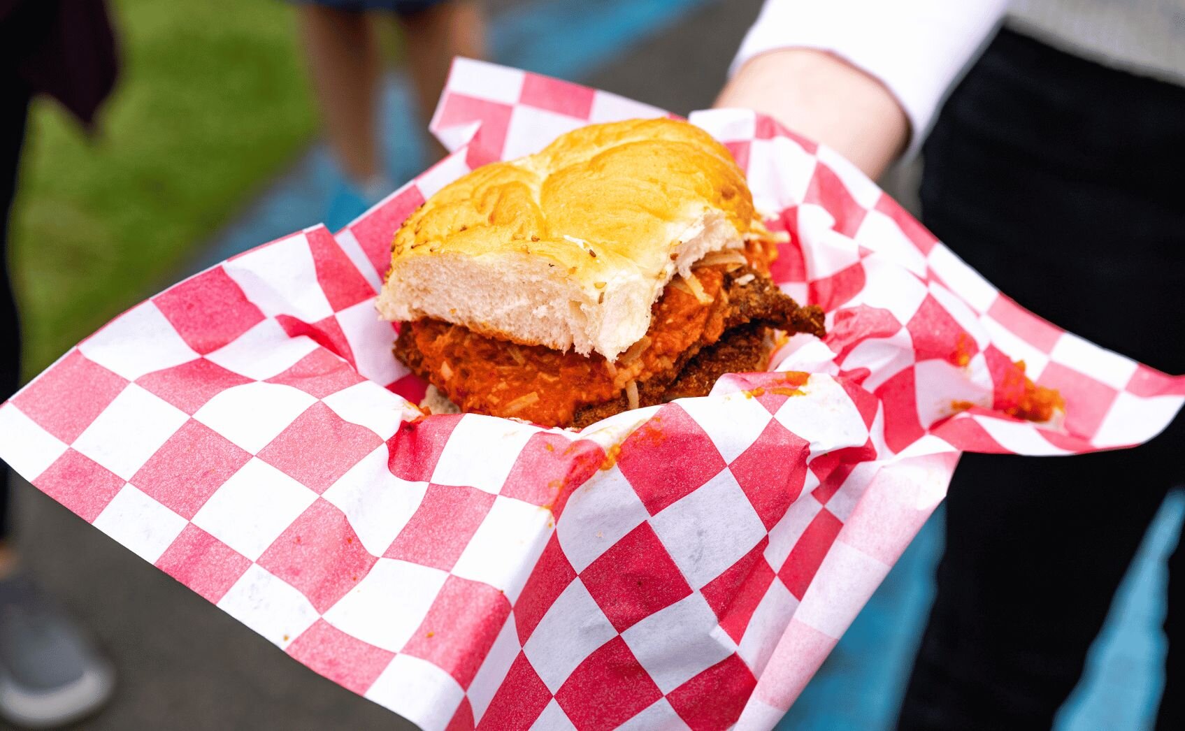 Outdoor food festival with people sitting at orange plastic chairs and wooden tables on artificial turf, food trucks visible
