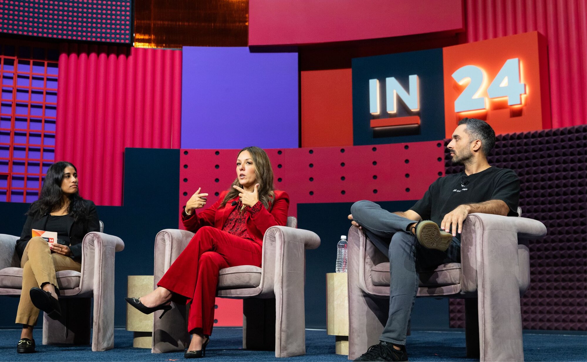 Three people sitting in gray chairs on a panel discussion stage with colorful geometric backdrop featuring 'IN24' logo