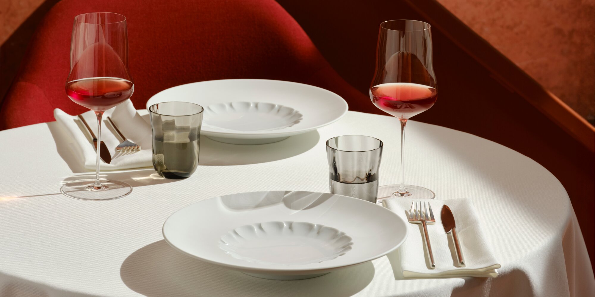 "Elegant gedeckter Tisch mit weißem Porzellan-Geschirr, filigranen Weingläsern mit Roséwein und rauchfarbenen Wassergläsern, arrangiert auf einer weißen Tischdecke in einem stilvollen Ambiente mit warmen Rottönen."
