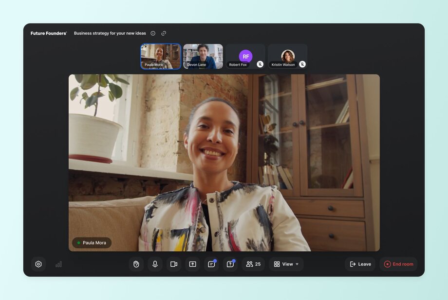 Live video meeting interface with speaker Paula Mora and participant thumbnails, chat, and control icons at the bottom of the screen.