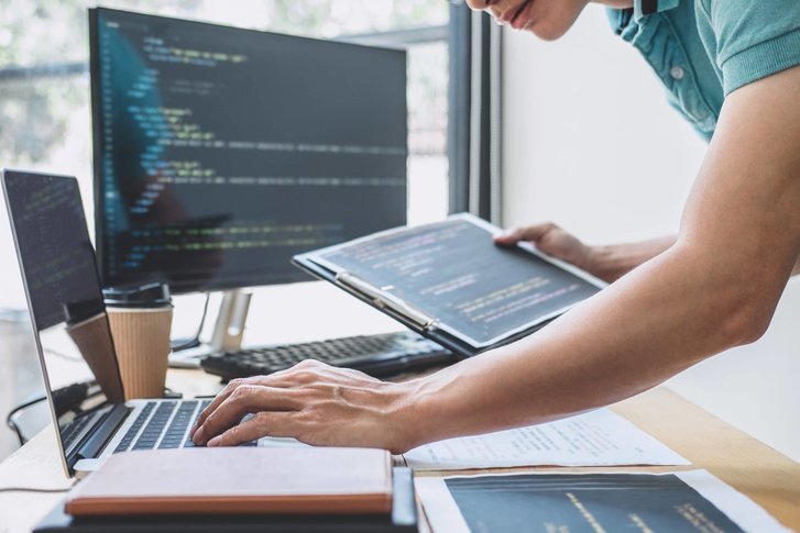 Person writing on a laptop with one hand and holding a folder with the other. A computer screen showing code on the background 