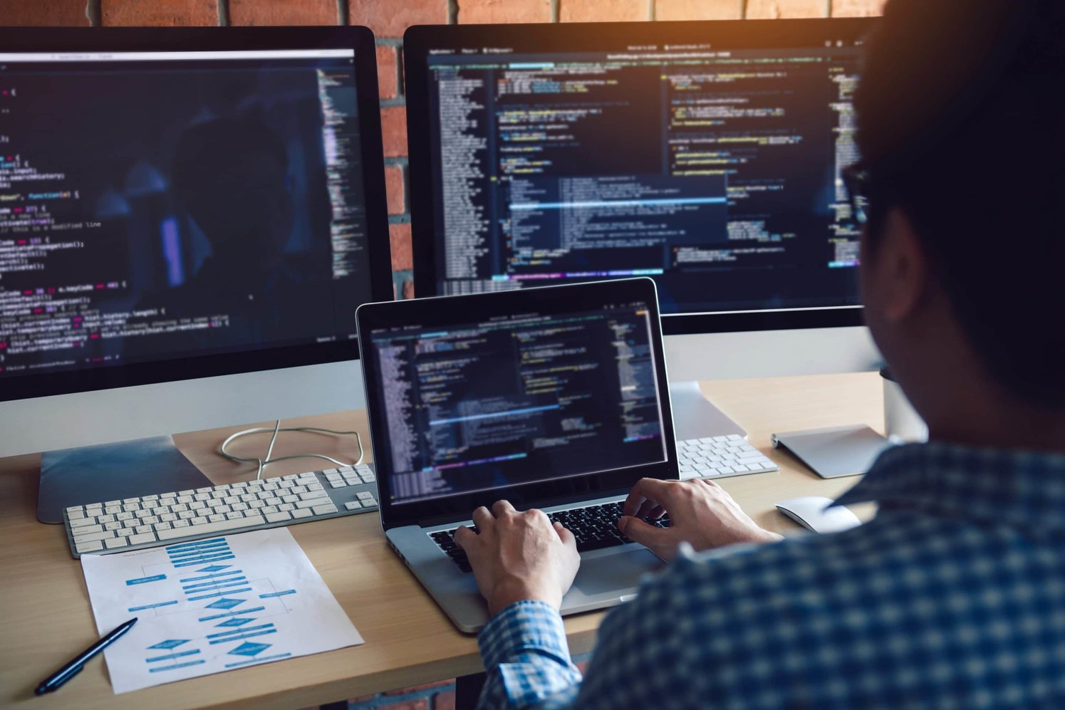 Man coding on his laptop with two other computer screens behind the laptop displaying code