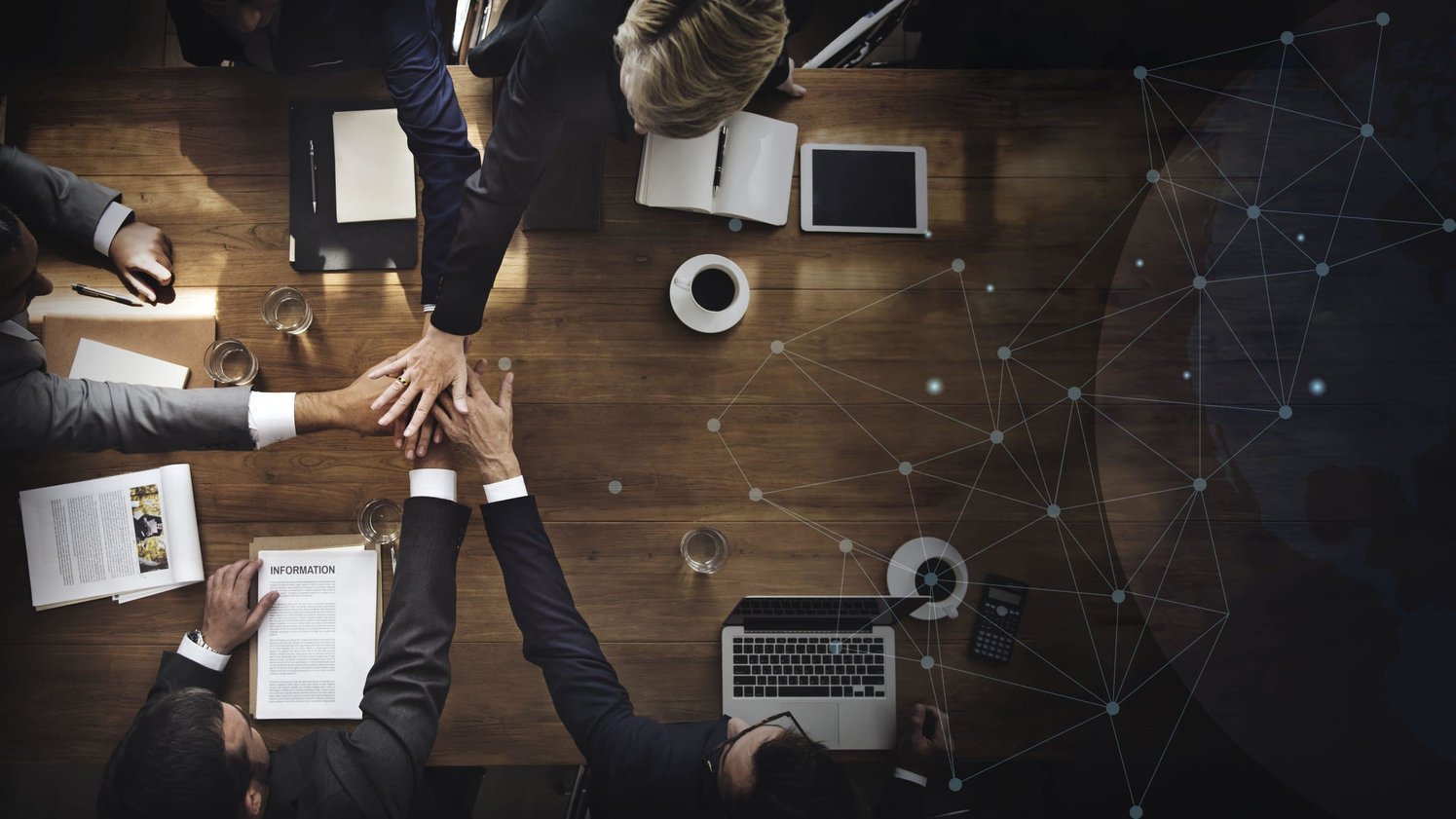 Three people putting their hands over other on the center, there are coffee and papers around them. 