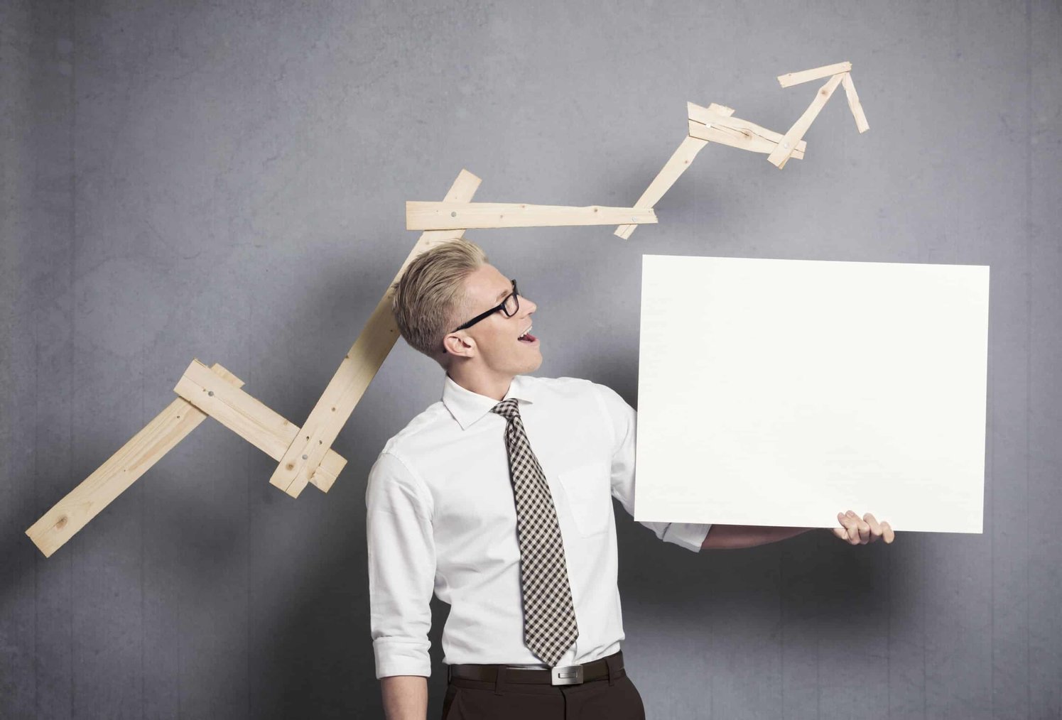 The man is holding a white cardboard, and behind him, a graph showing an upwards trend made of wood. 