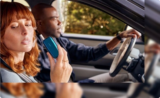 A woman is smoking from the Ploom device in a car.