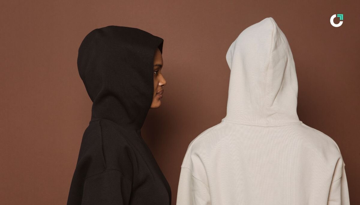 Two people standing in front of a brown background, one in a black hoodie and the other in a white hoodie