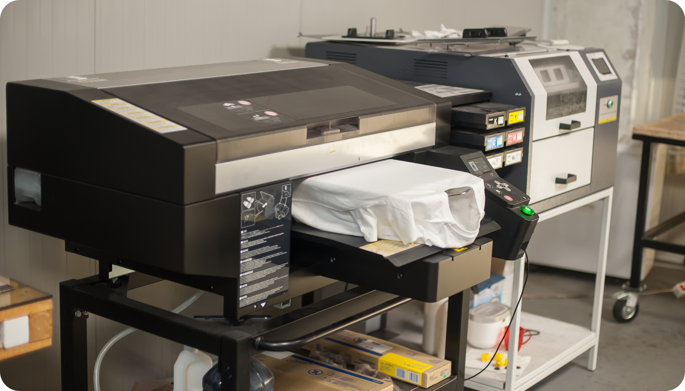 A direct-to-garment (DTG) printer printing a white t-shirt, with visible ink cartridges and control panel in a production workspace.