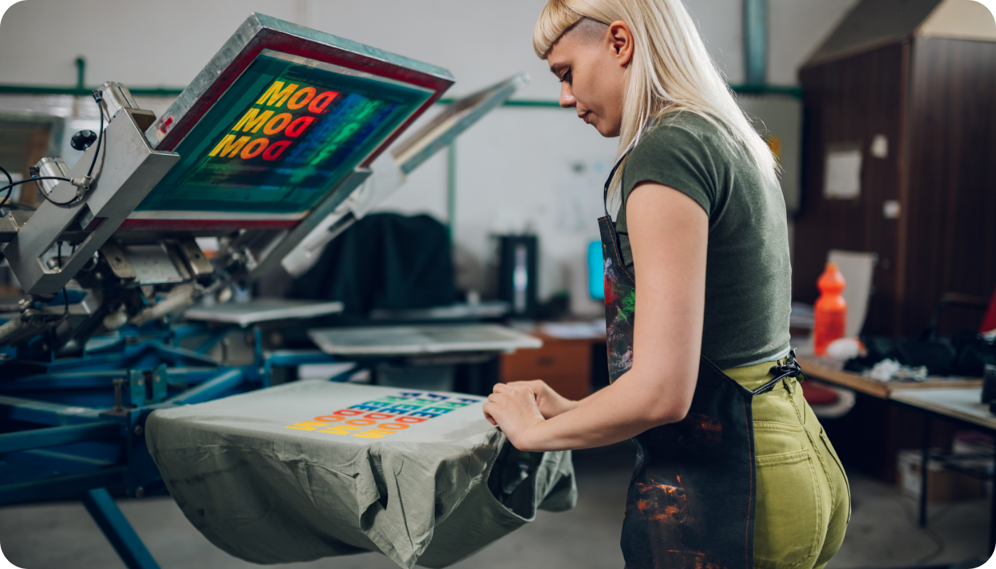 A woman operating a screen printing press, preparing to transfer a colorful multi-layered design onto a t-shirt, demonstrating traditional and highly durable t-shirt printing method used for bulk production.