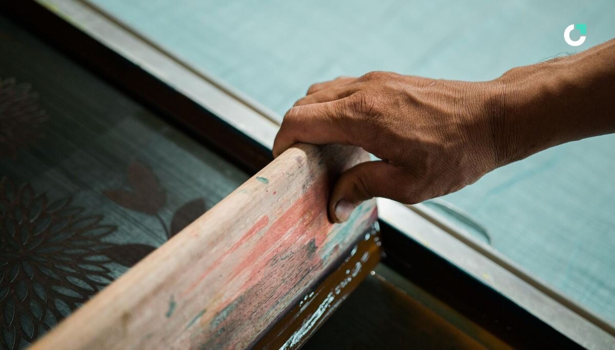 Hand in the corner of the photo holds a squeegee, halfway through screen printing a design.