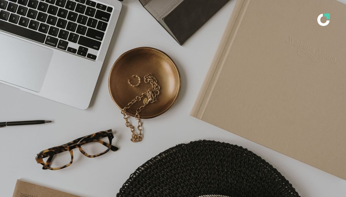 Flat lay of a creative workspace with a laptop, tortoiseshell glasses, gold jewelry, and neutral-toned accessories — evoking a refined aesthetic ideal for brainstorming elevated, design-forward merch ideas.
