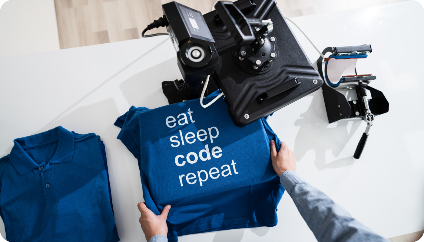 Top-down view of a person placing a blue t-shirt under a heat press machine with the printed text “eat sleep code repeat,” showing the heat transfer printing process in action.