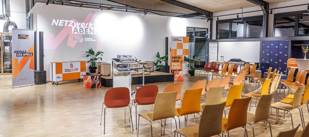 Mehrere Reihen unterschiedlich farbiger Stühle vor einer Bühne mit Bannern und einem Tisch in einem großen Raum mit Holzboden und sichtbaren Deckenrohren