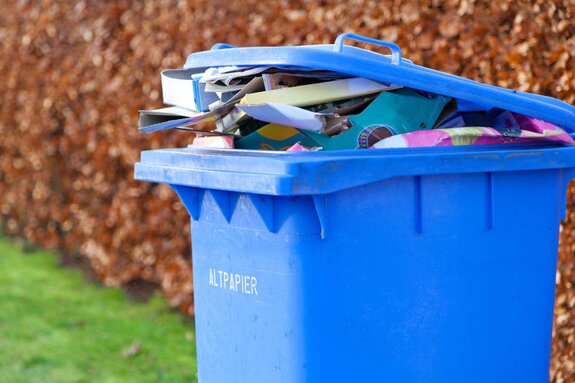 Blaue Altpapiertonne mit offenem Deckel, gefüllt mit Papierabfällen, im Freien vor einer Hecke