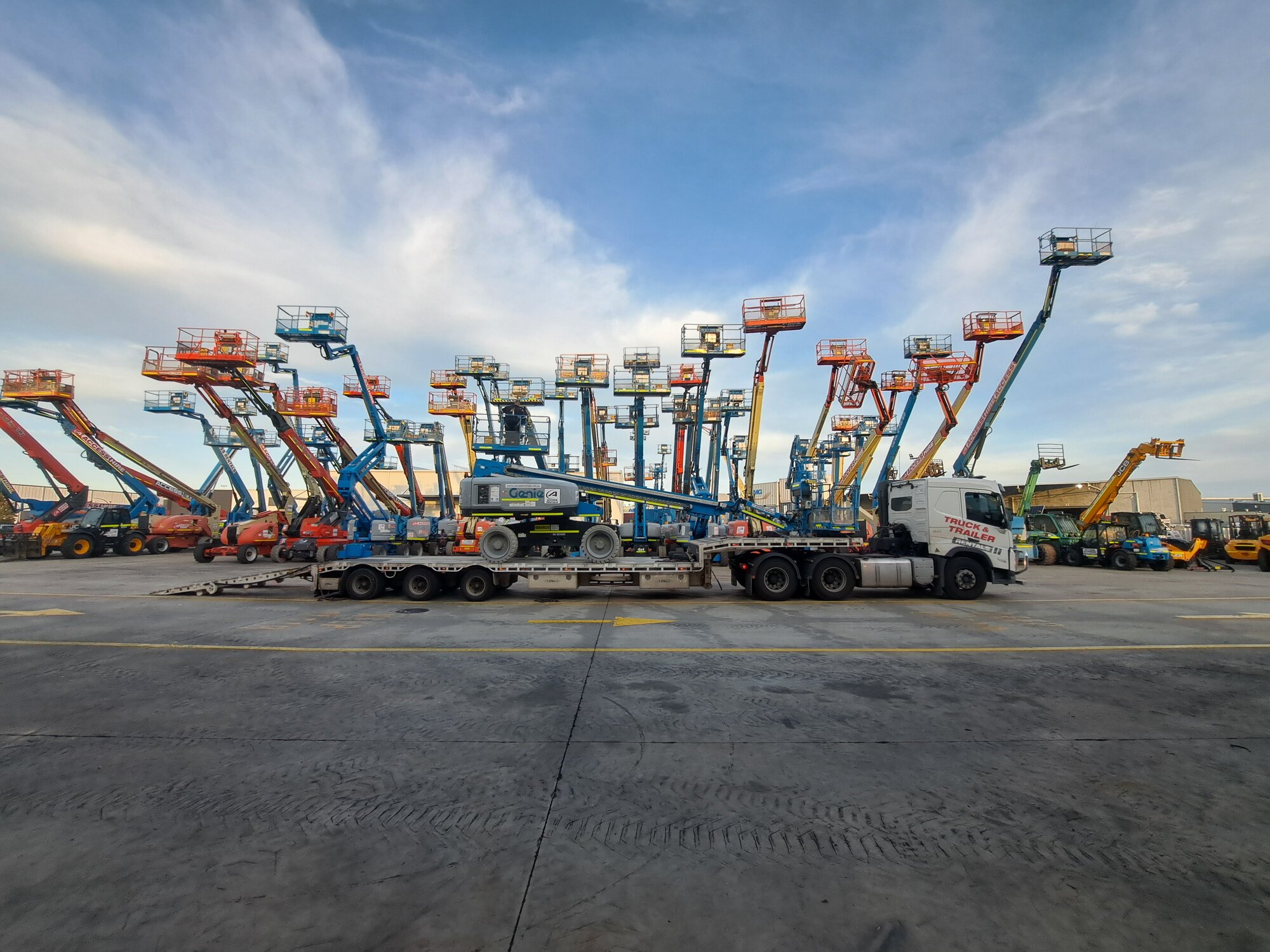 Truck transporting a Genie boom lift through the Access Hire yard in Canning Vale, Western Australia, with the access equipment fleet in the background.