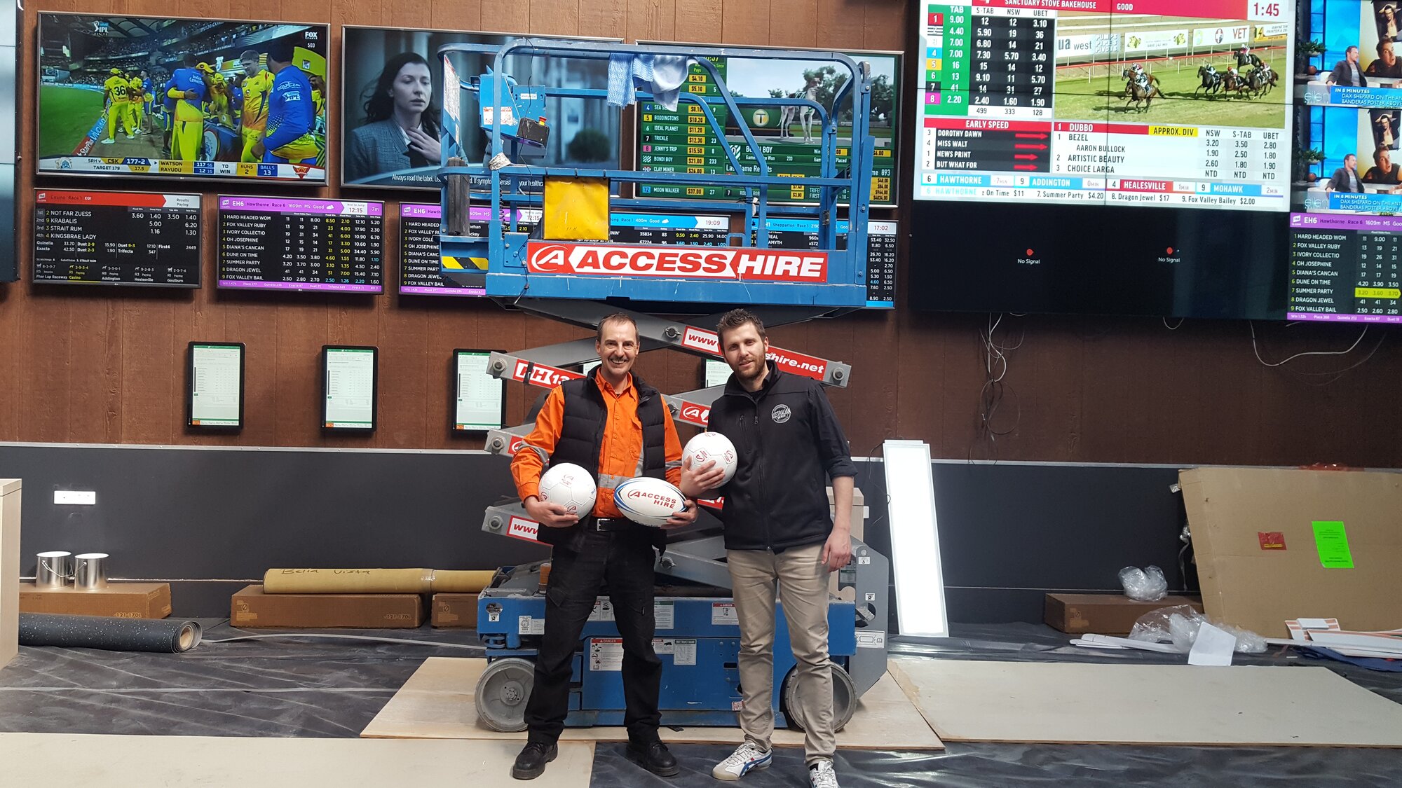 Two people holding Access Hire branded footballs in front of an Access Hire scissor lift at a commercial venue in Sydney.