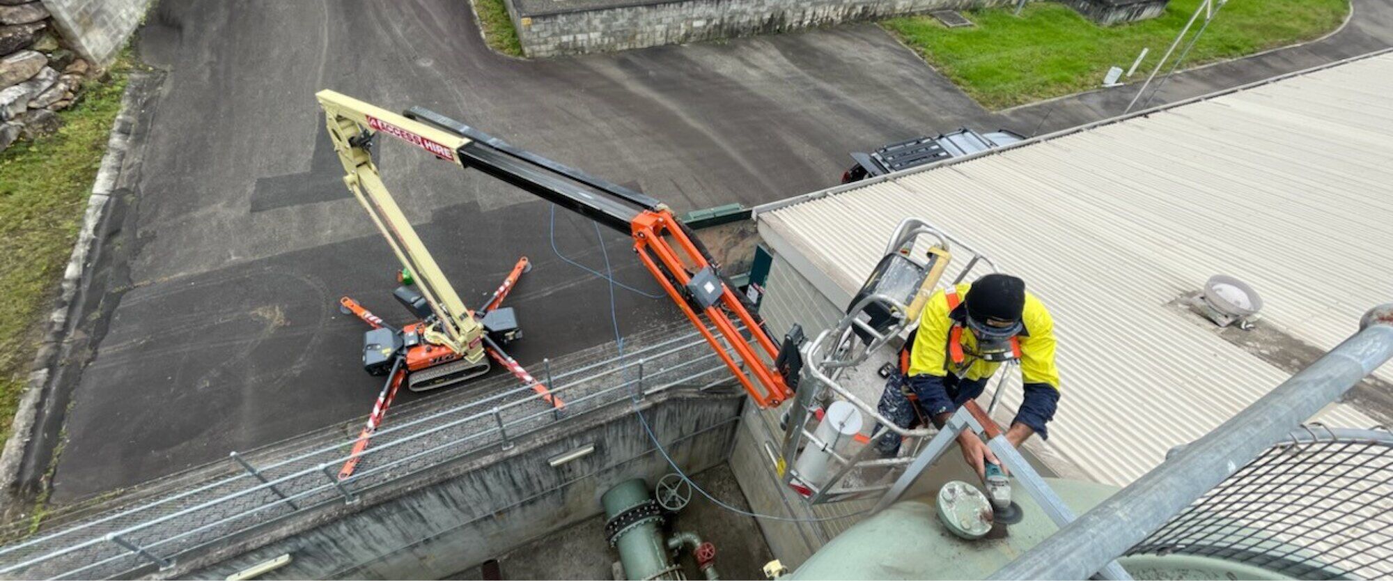 Worker using a tracked articulating boom lift to perform maintenance on industrial equipment at height, with the lift positioned beside a building.