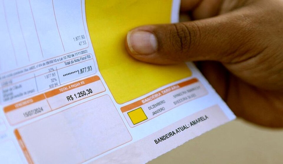 Imagem de uma mão segurando uma conta de luz. Na conta uma indicação de bandeira amarela na conta de luz.
