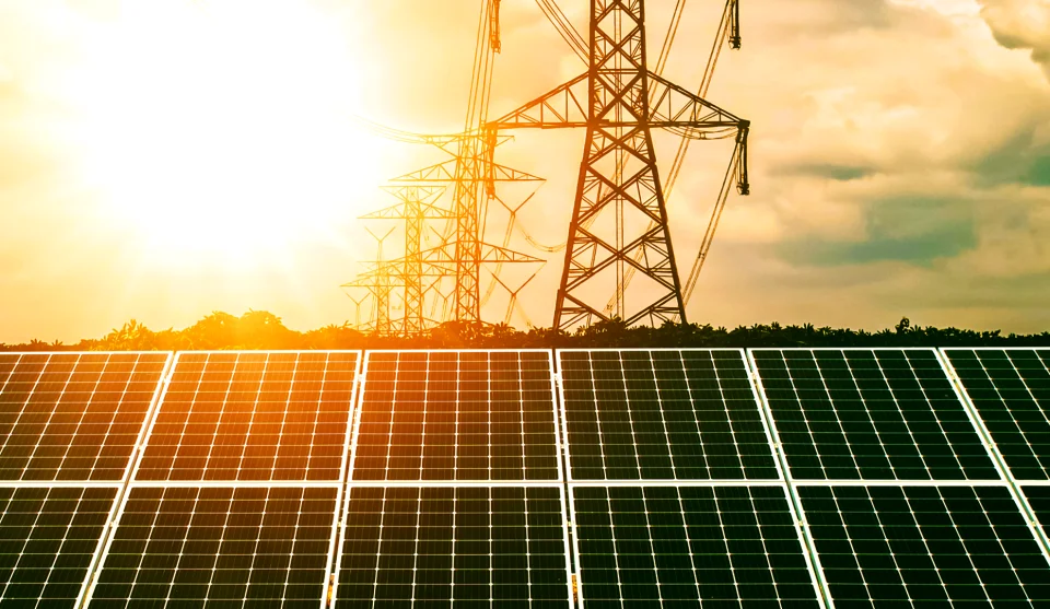 Fotografia de um horizonte com uma usina solar a frente e ao fundo uma estrutura de distribuidoras elétricas e mais ao fundo um sol radiante.
