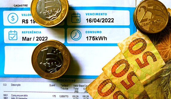 Fotografia de uma conta de luz com zoom em seus detalhes, em cima da conta de luz algumas moedas de 1 real, 25 centavos e notas de 50 reais.