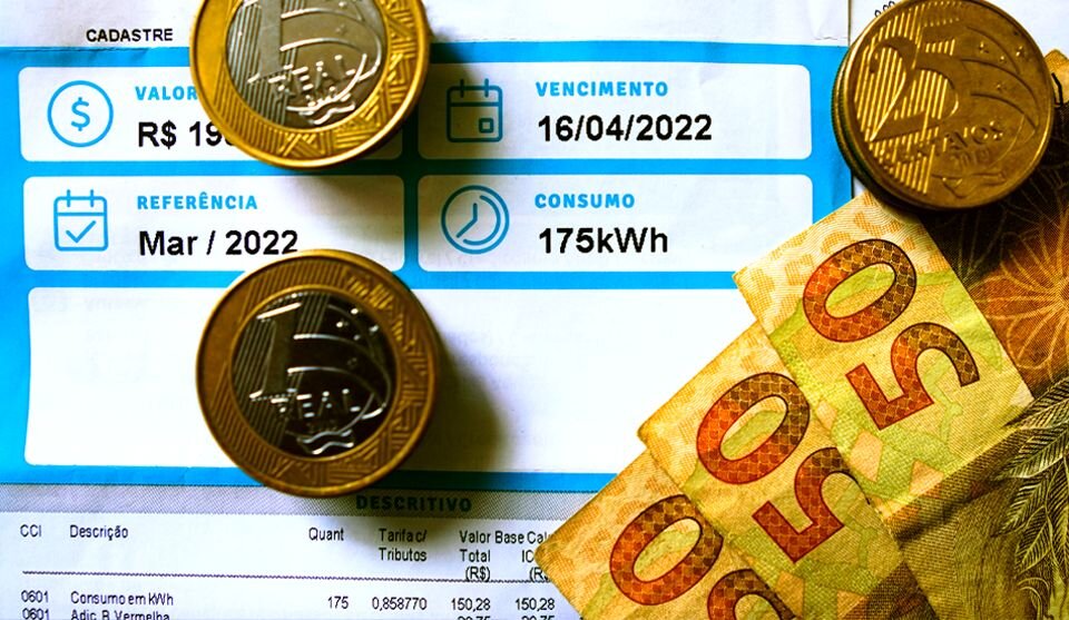 Fotografia de uma conta de luz com zoom em seus detalhes, em cima da conta de luz algumas moedas de 1 real, 25 centavos e notas de 50 reais.