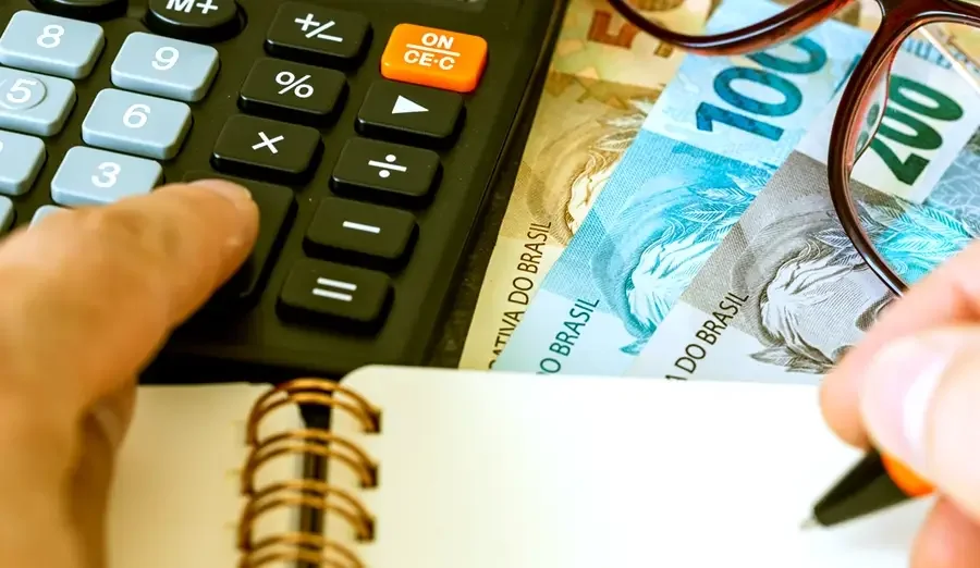 Foto focada em uma mão mexendo em uma calculadora e a outra anotando com caneta em um caderno, ao fundo notas de reais brasileiros e um óculos em cima das notas.
