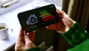 Fotografia de uma mão de uma mulher idosa segurando um celular em suas mãos, na tela do celular um indicativo de gasto com energia na casa, ao fundo da imagem um parapeito de janela iluminada pelo luz do dia e com uma caneca de café em cima.