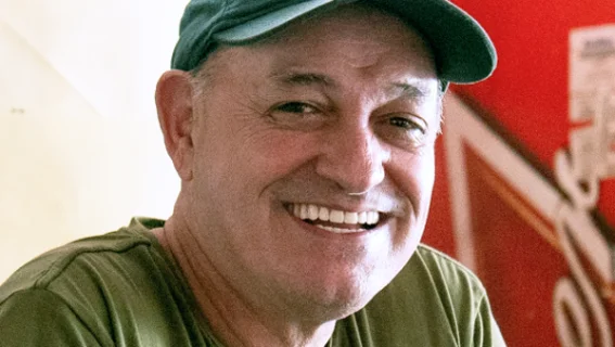 Ildefonso: Homem em frente às geladeiras de um bar, sorrindo para a câmera, vestindo camisa e boné verdes.